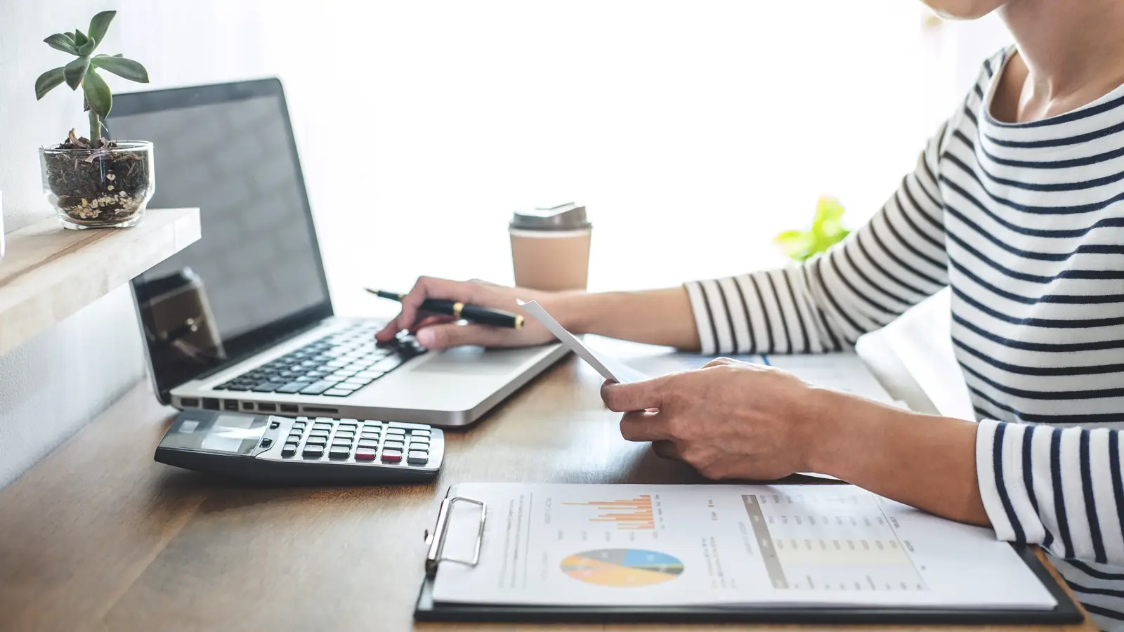 Person working on accounts on a laptop with charts on paper